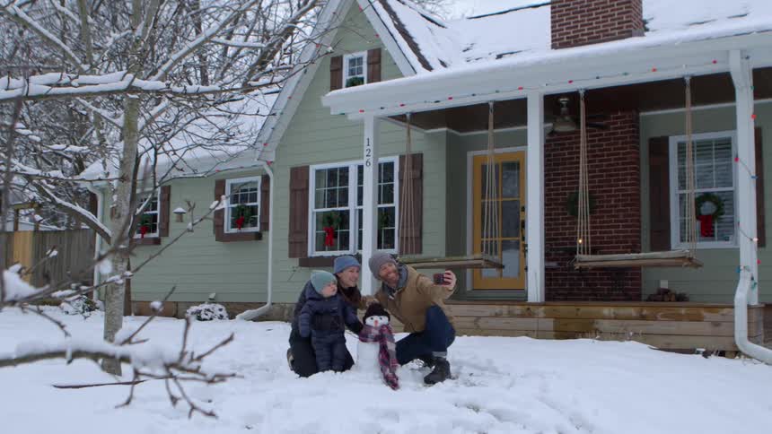 Two parents and a young child take a selfie near a small snowman in their yard