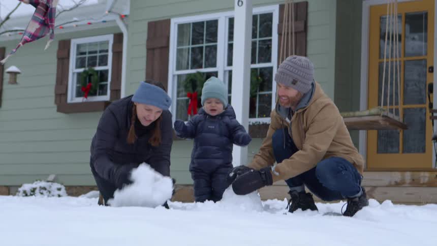 A family builds a small snowman in front of a house with a green exterior and a porch