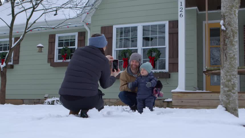 A woman taking pictures of a man who is crouching next to his little boy and they are in a snowy environment in front of a house