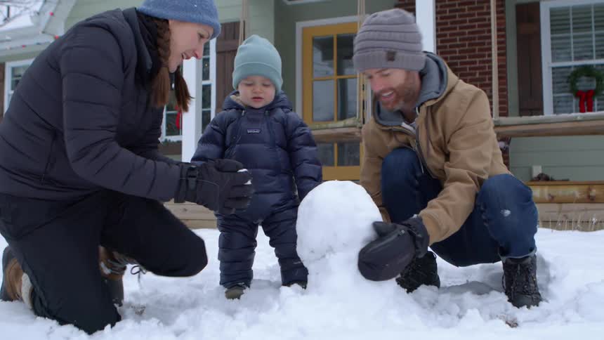 Parents show their child how to make a snowman in their front yard on a winter day