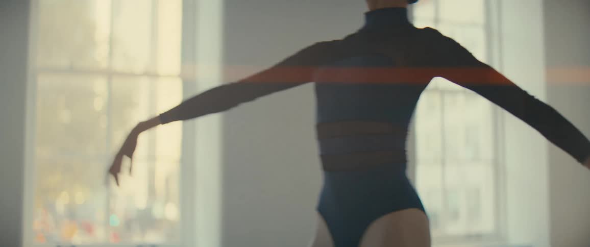 A woman performing a ballet pose with arms extended in a well lit room with large windows in the background