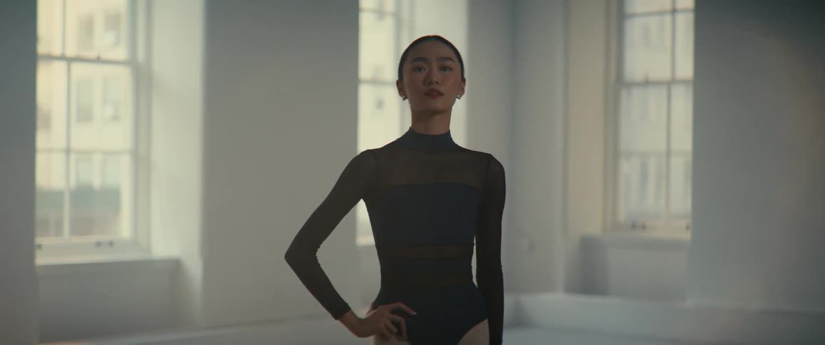 An asian ballerina stands in a dance studio with her hand on her hip