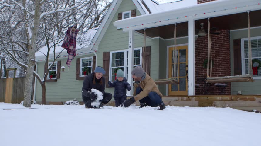 A family builds a small snowman in front of a house with a green exterior and a porch