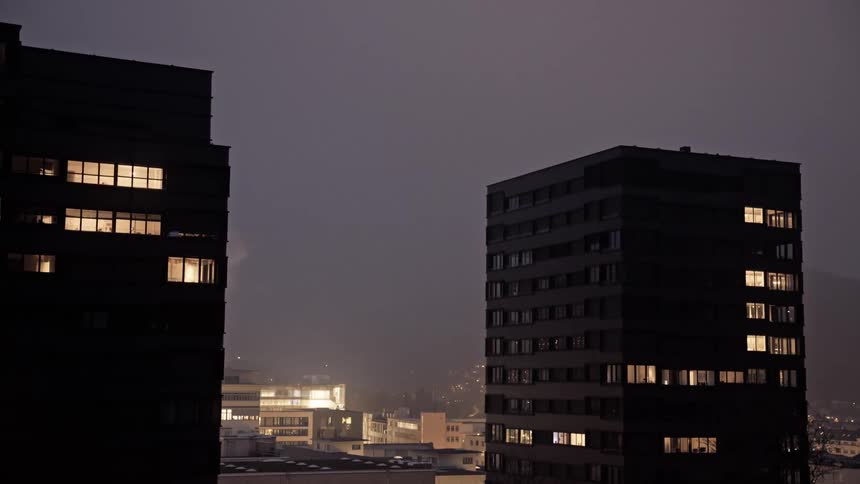 Two tall blocky buildings with a few illuminated windows stand against a dark early evening sky with a hazy cityscape