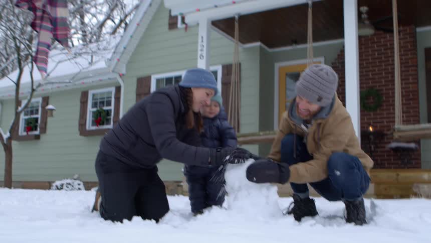 Father and mother next to their little son building a snowman in the backyard of a house