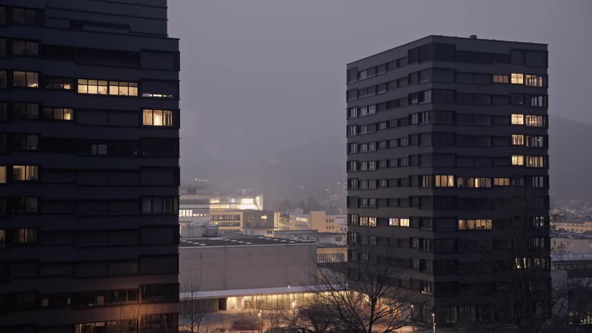 Two tall modern buildings with illuminated windows stand prominently against a dark early evening sky overlooking a cityscape with blurred lights