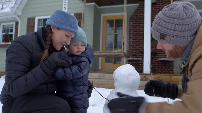 A young family builds a snowman in front of their house at christmastime