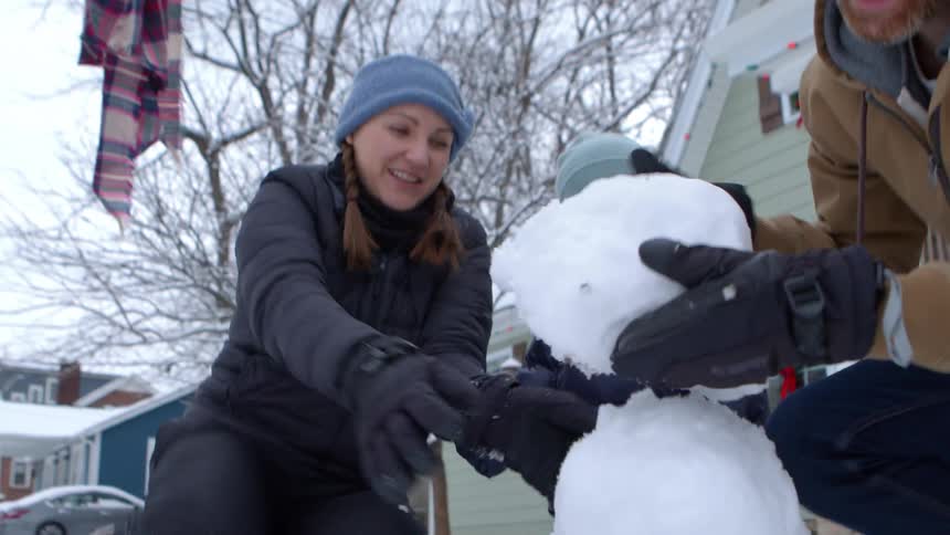 A family builds a small snowman in front of a house with a green exterior and a porch