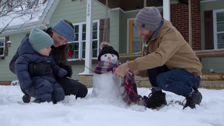 A family builds a small snowman in front of a house with a green exterior and a porch the snowman is wearing a hat and scarf