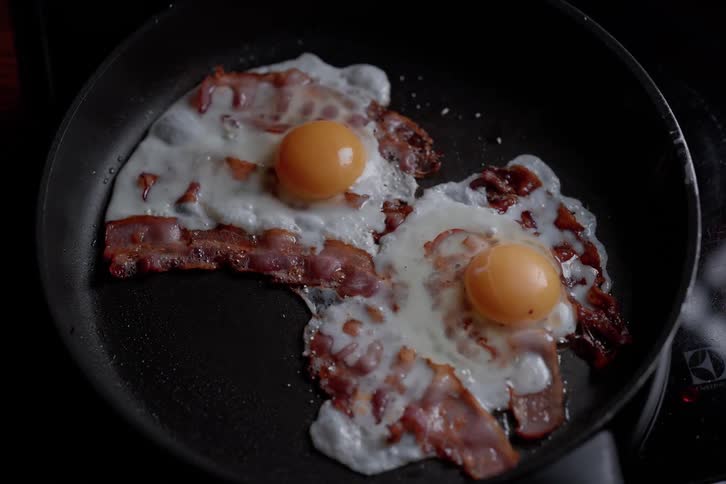 Two sunny side up eggs nestle amongst crispy curled pieces of bacon in a dark nonstick skillet their golden yolks bright against the white of the eggs and the reddish brown bacon