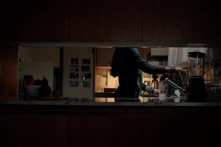 A person is seen from behind through a kitchen passthrough window drinking a glass of water at the kitchen sink