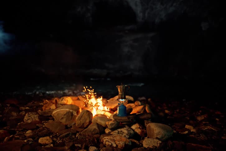 Smoke and sparks rise from a riverside campfire while a kettle heats on a portable burner at night