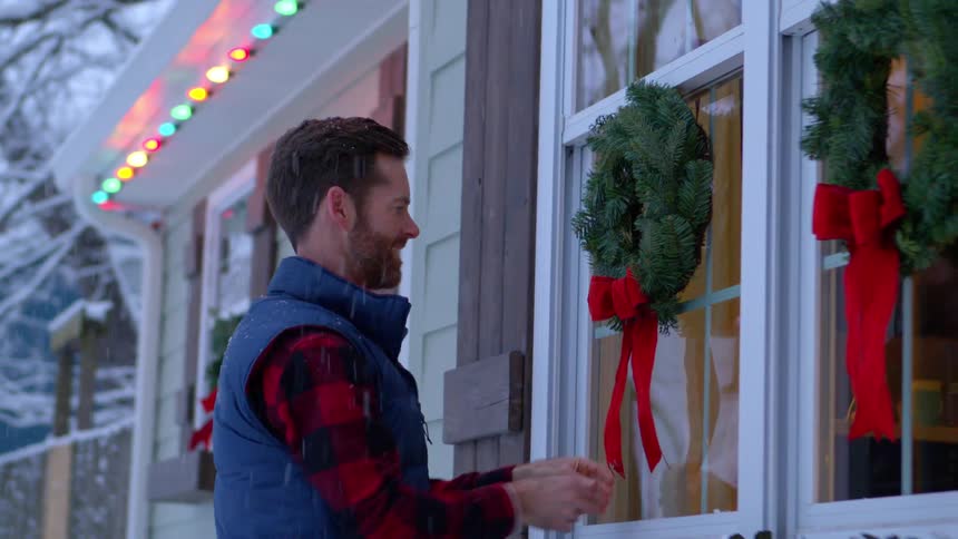 Smiling man hangs a wreath on his window in the lightly falling snow
