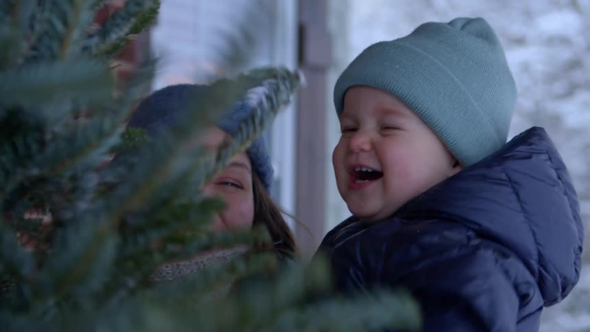 A little boy on a womans lap is hitting a pine tree leaf happily and they are wearing winter clothes