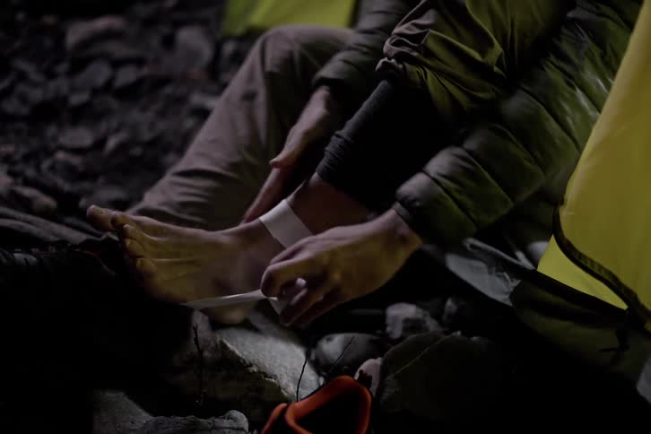 A person sitting at the entrance of a tent wraps a bandage around their foot on rocky ground with one shoe removed and placed nearby