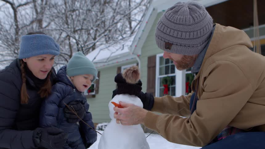A family builds a small snowman in front of a house with a green exterior and a porch