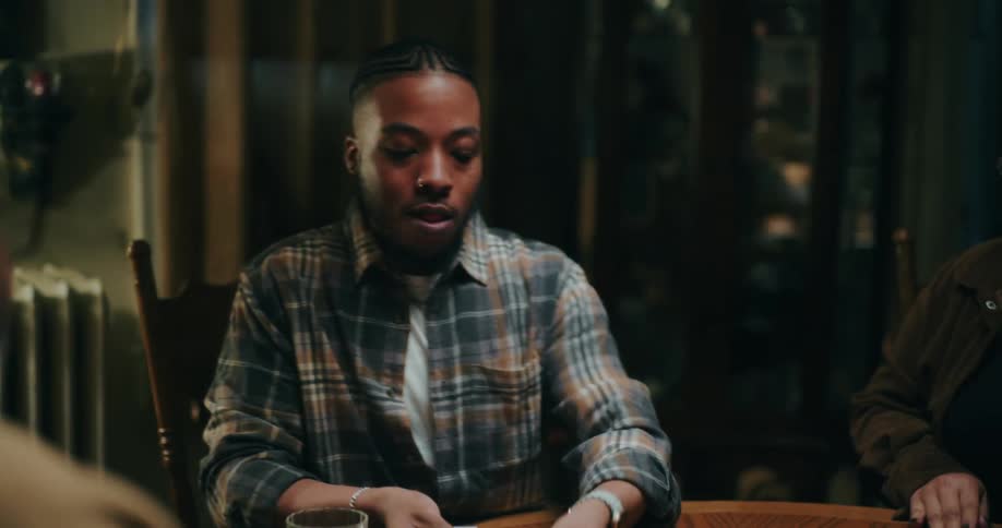 A young man with braided hair and a nose piercing wearing a plaid shirt is playing cards at a wooden table