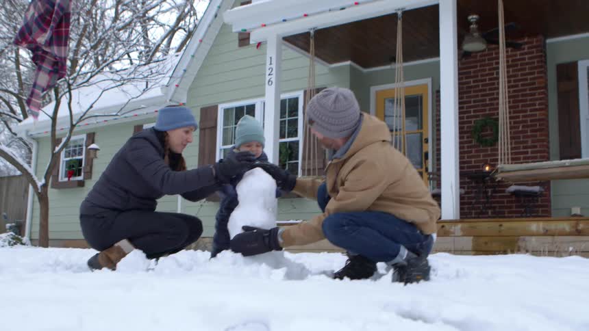 A family builds a small snowman in front of a house with a green exterior and a porch
