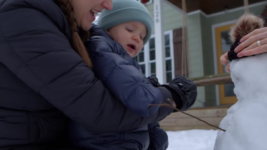 A mother and father build a snowman with their toddler son in front of their home