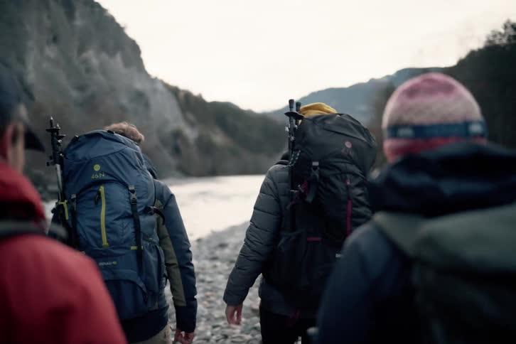 Four backpackers are walking beside a river in a mountain range on a cloudy winter day