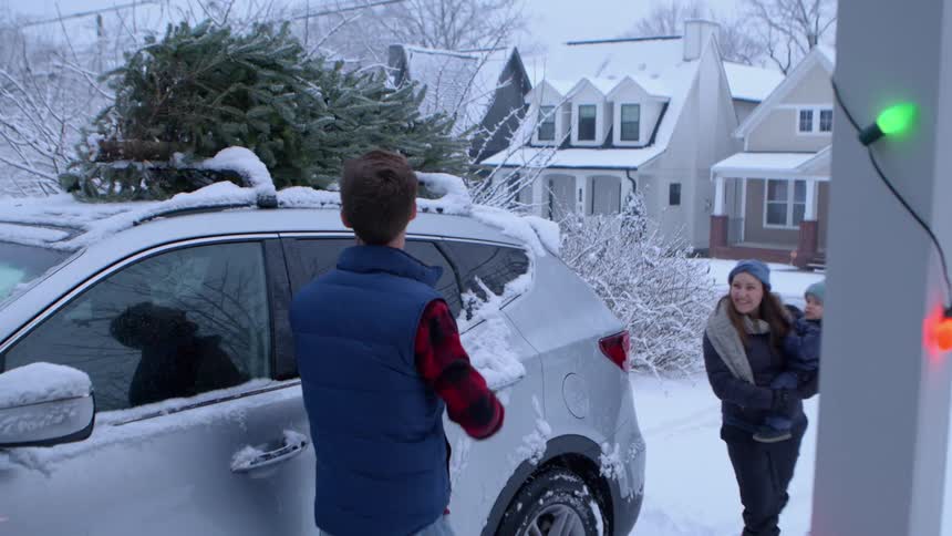 A man unloads a christmas tree from the roof of a snowcovered car while his wife holds their son with houses and holiday lights in the background
