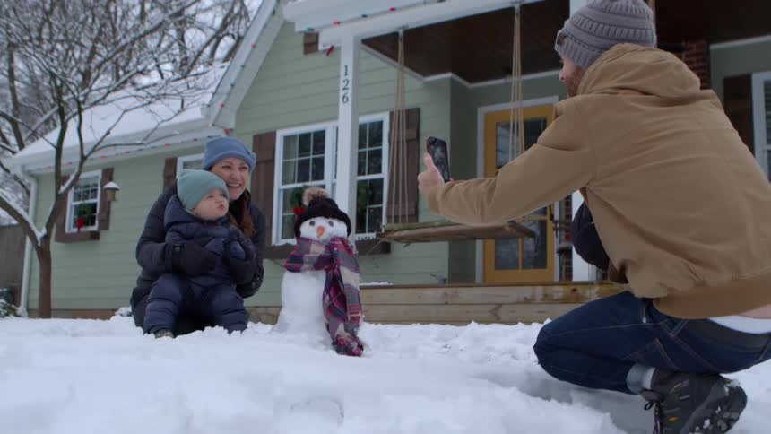 A man taking pictures of a woman hugging a little baby who is next to a snowman in front of a house in winter with snow on the ground