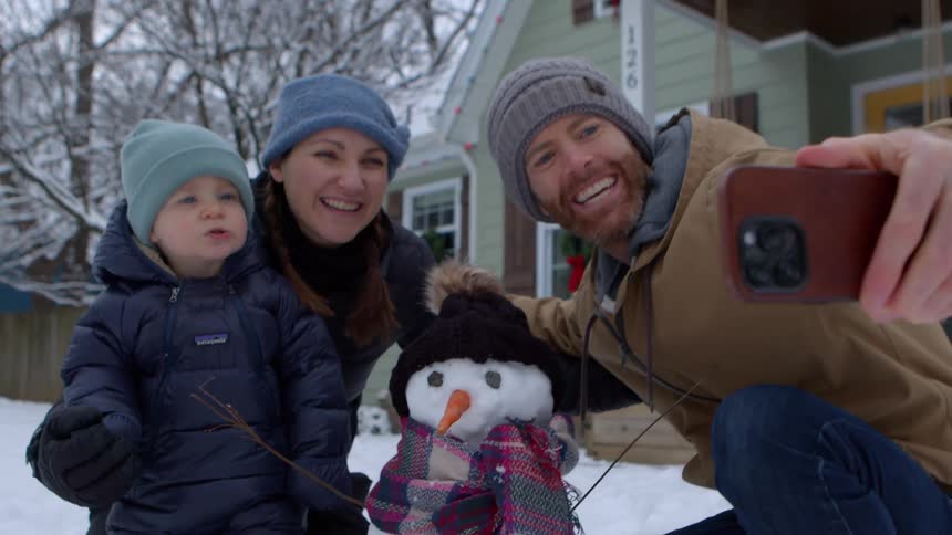 A family takes a selfie with a small snowman in front of a house during winter