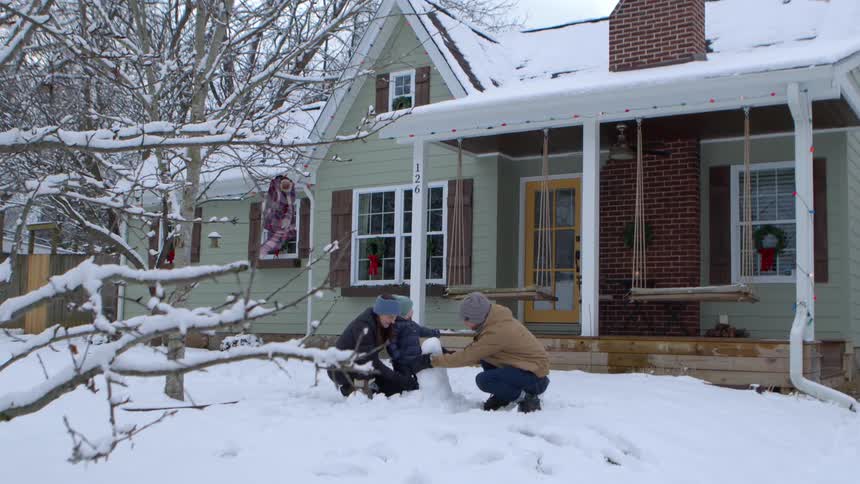 A family builds a small snowman in front of a house with a green exterior and a porch