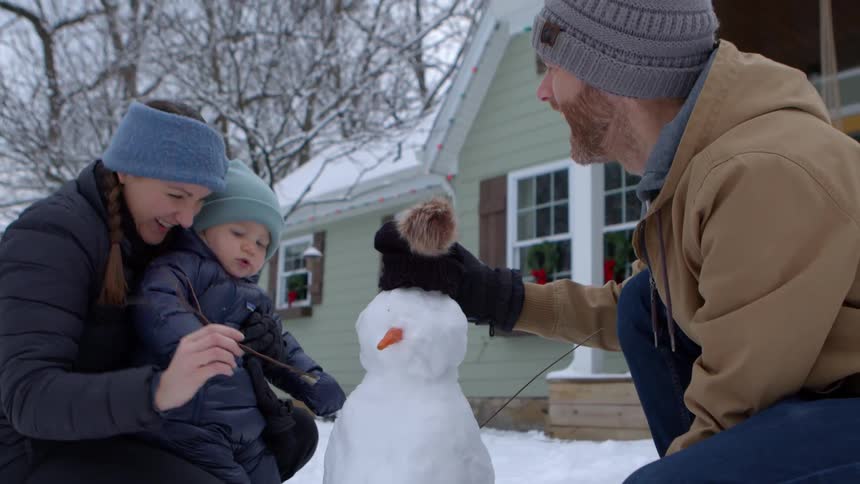 Father and mother with their little son building a snowman in the backyard of a house