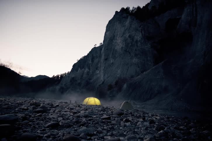 A litup tent glows yellow in the fading light with the shadow of a person moving inside projected onto the tent wall as the camera steadily approaches across the rocky ground in a secluded mountain setting