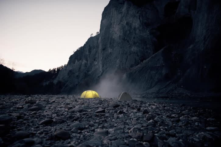 A brightly lit yellow tent sits on a rocky ground near a large dark rock formation against a twilight sky