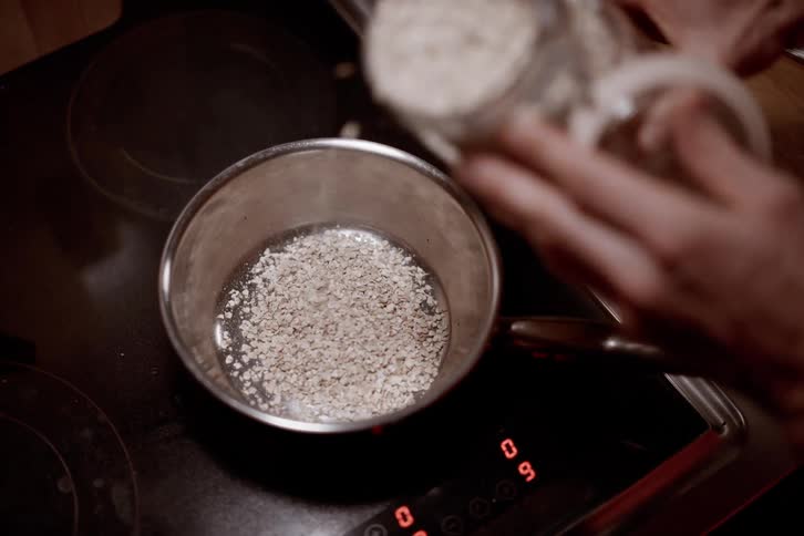 Pair of hands pouring oatmeal powder into a pot on the stove