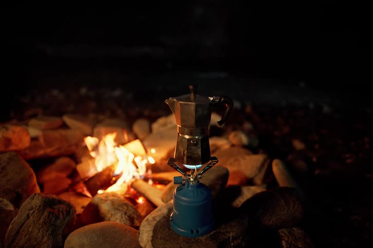 Aluminum percolator heating on a portable butane canister next to a campfire at night