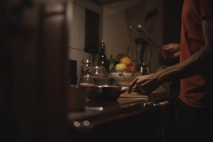 A person grates a fruit over a pan on a stovetop in a dimly lit kitchen