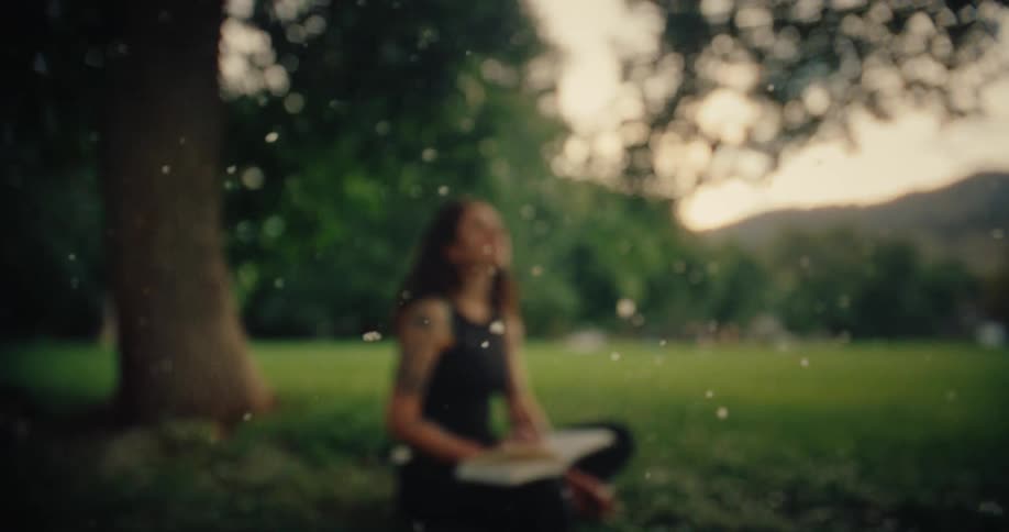 A woman with long hair and tattoos on her left arm sits outdoors under a tree with a book on her lap surrounded by floating white particles