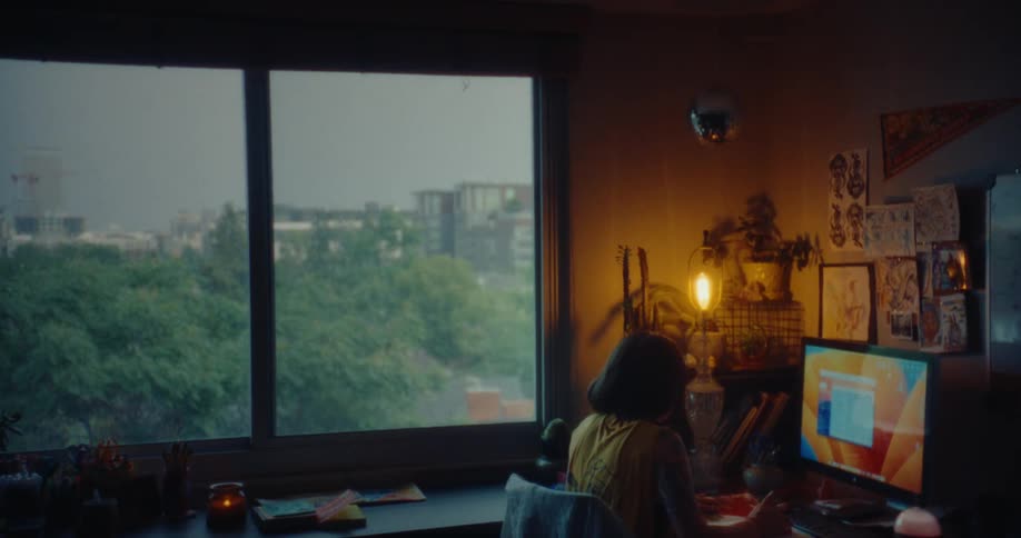 A woman is sitting at a desk in a dimly lit room working on a computer with a large window showing a view of trees and buildings outside
