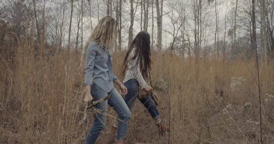 Women walk together with campfire supplies through tall grass