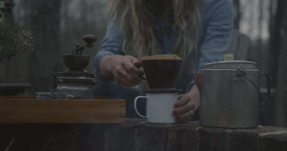 A woman makes coffee next to a campfire