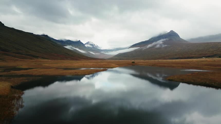 A still dark lake reflects the cloudy sky and surrounding brown and gray mountains with a small green house visible in the distance