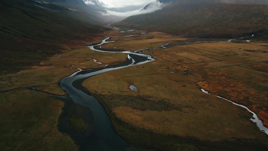 A winding river meanders through a vast brown and green valley framed by mountains partially shrouded in mist under an overcast sky