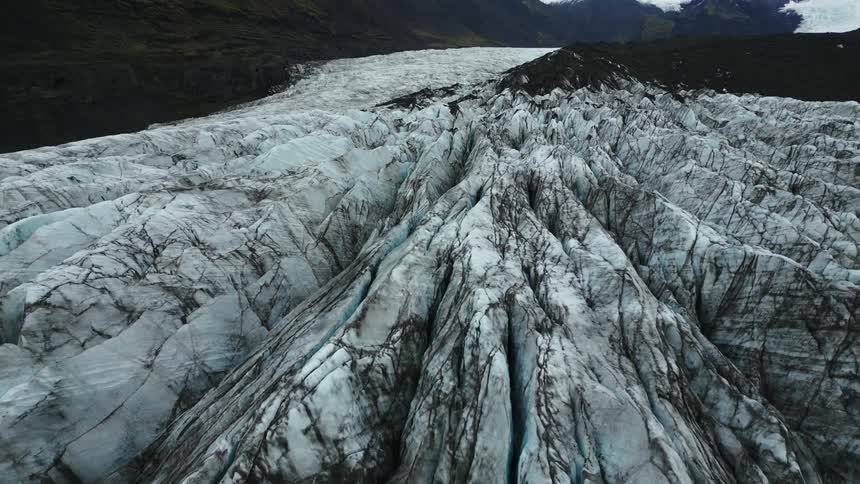 A textured glacier surface stretches out revealing a pattern of cracks and crevices in shades of gray and white with dark rock visible at the top