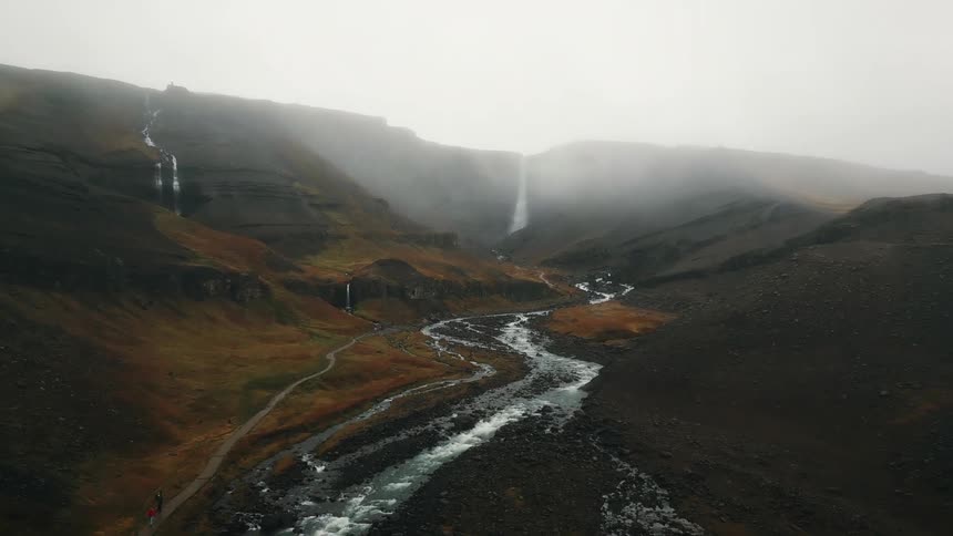 An aerial view of a river with several pebbles on the sides and in front of a mountain with a thick fog on top