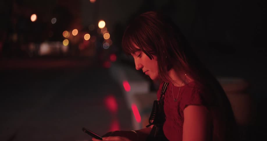 The face of a woman who is using her cell phone in a dark outdoor environment with some blurred lights in the background
