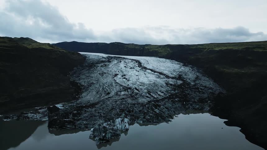 A glacial tongue stretches down from rugged mountains its textured surface reflecting the cloudy sky above a dark still lake