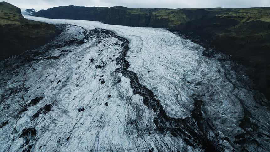 A glacier with a prominent white ice surface streaked with dark debris flows between two dark rocky mountain ridges under an overcast sky