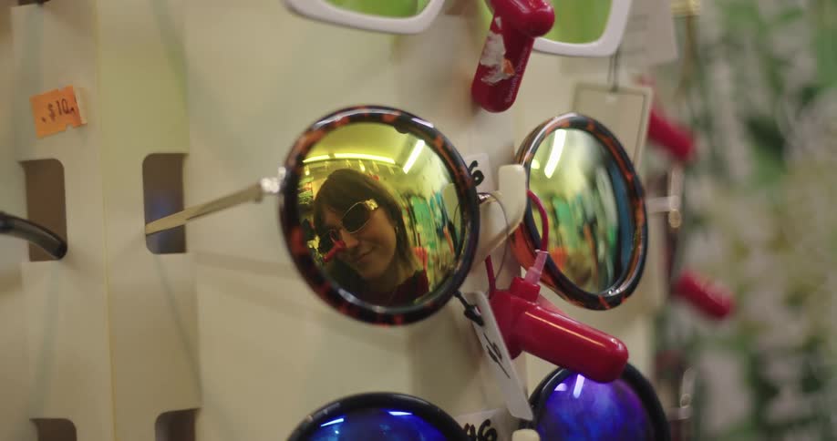 Close up of a pair of sunglasses on a display case reflecting the face of a woman trying on another pair of sunglasses