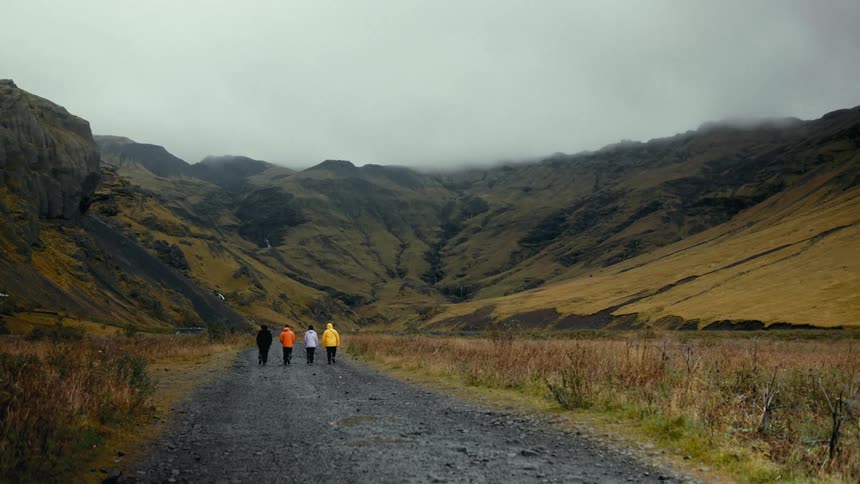 Four people are walking on a gravel path through a misty mountainous landscape with grassy slopes and rugged terrain