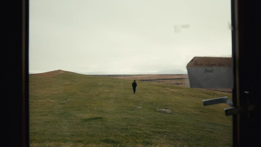 A person is walking alone on a vast grassy field towards a distant hill under an overcast sky viewed from inside a house