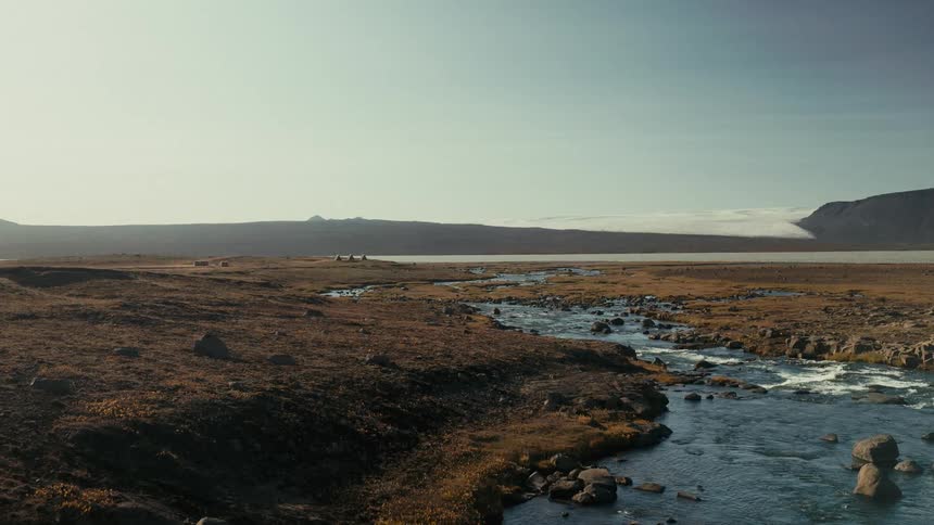 A wide shot shows a river flowing through a grassy landscape with rocks distant mountains and a bright sky