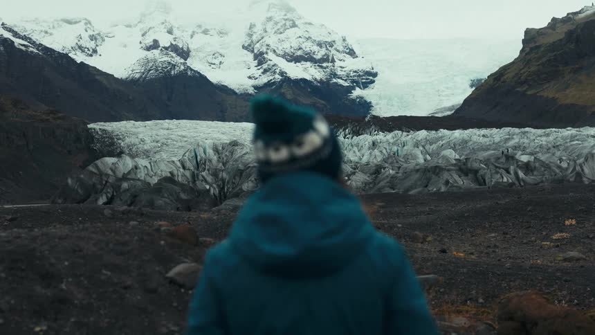 A person in a blue jacket and beanie stands on rocky terrain facing a vast glacier with snowcapped mountains in the background